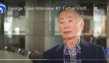 George Takei in a plaid collared shirt and jacket sits at a table for an interview